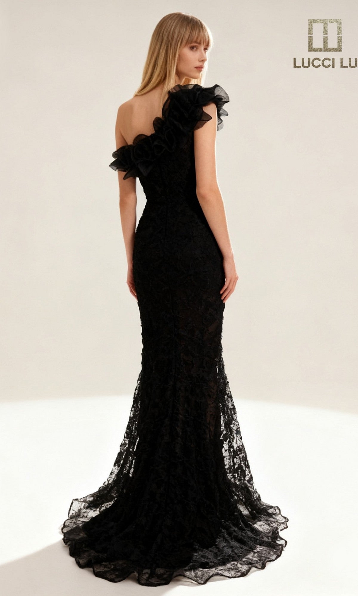A woman stands facing away in a brightly lit studio, wearing the Lucci Lu 6062 long prom dress—black, one-shoulder, with lace, ruffle detail, and a fitted silhouette.